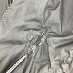 May include: A dark gray fabric surface featuring a jagged, irregular tear in the material. The edges of the rip are frayed, revealing a darker, textured layer underneath the top layer of the fabric. A metallic pen tip points toward the damaged area.