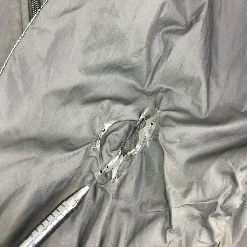 May include: A dark gray fabric surface featuring a jagged, irregular tear in the material. The edges of the rip are frayed, revealing a darker, textured layer underneath the top layer of the fabric. A metallic pen tip points toward the damaged area.