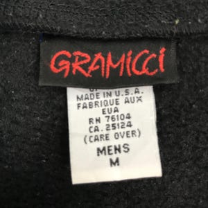 以下が含まれることがあります： 赤色の「GRAMICCI」ロゴが入った黒い衣類のタグのクローズアップ。ロゴの下には、「MADE IN U.S.A.」、「MENS」、サイズ「M」などのテキストが表示された白いタグがあります。タグは黒い生地に付いています。