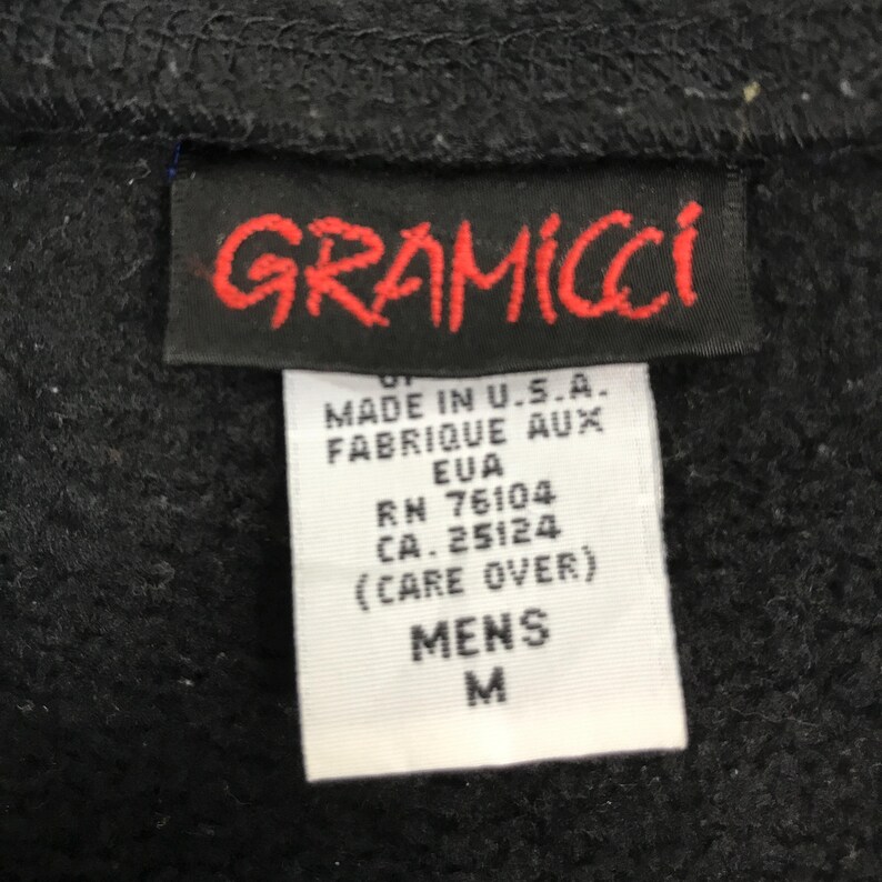 以下が含まれることがあります： 赤色の「GRAMICCI」ロゴが入った黒い衣類のタグのクローズアップ。ロゴの下には、「MADE IN U.S.A.」、「MENS」、サイズ「M」などのテキストが表示された白いタグがあります。タグは黒い生地に付いています。