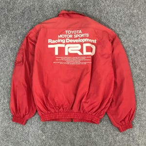 May include: Red Toyota Motor Sports Racing Development jacket with white text and logo. The jacket has a collared neckline, elastic cuffs, and a zippered front. The back of the jacket features the TRD logo and additional text.