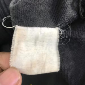 May include: A rectangular white fabric garment care label with horizontal ribbed texture, stitched into the interior seam of a dark charcoal gray corduroy fabric garment. The label is blank and shows signs of fraying threads at the edges.