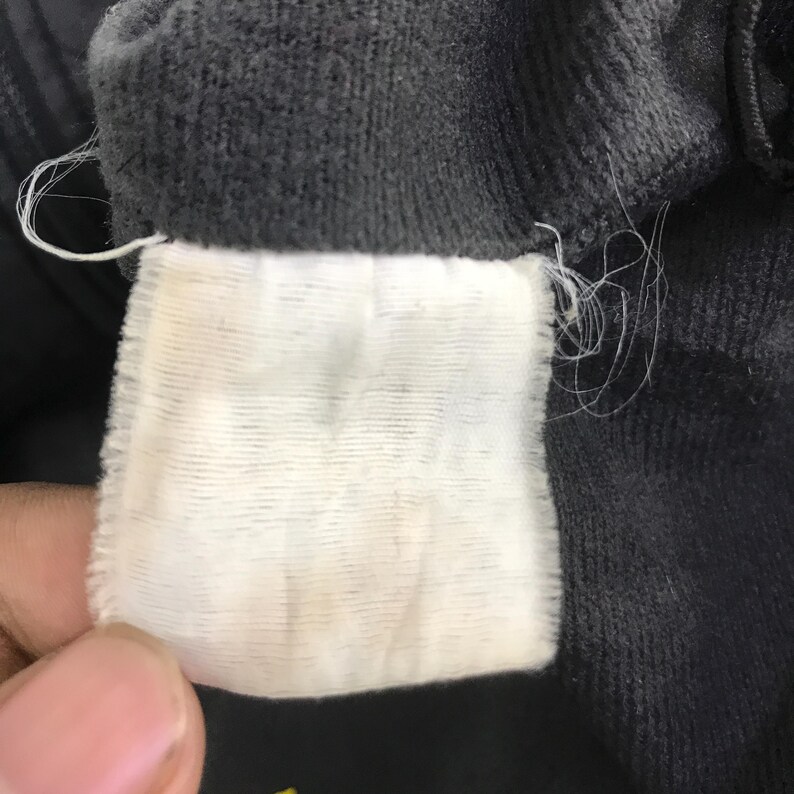 May include: A rectangular white fabric garment care label with horizontal ribbed texture, stitched into the interior seam of a dark charcoal gray corduroy fabric garment. The label is blank and shows signs of fraying threads at the edges.