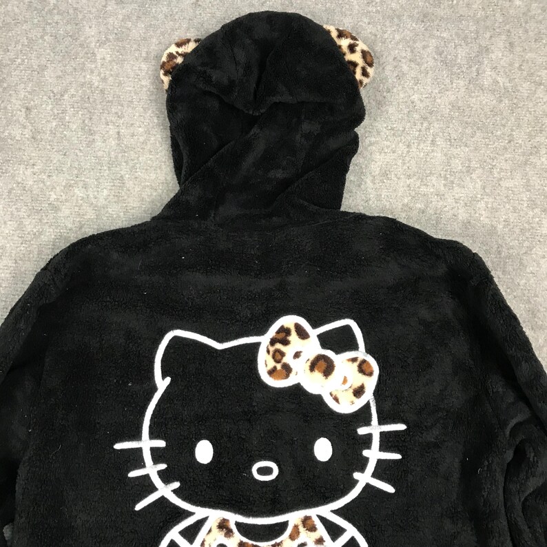 Pu&ograve; includere: Felpa con cappuccio in pile nero con grafica Hello Kitty bianca e orecchie e fiocco con stampa leopardata.