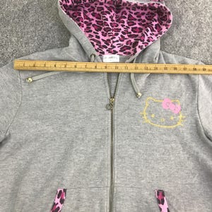 May include: A gray zip-up hoodie with a pink leopard print lining in the hood and pocket accents. The hoodie features a gold outline of Hello Kitty with a pink bow on the front. The zipper is gold.