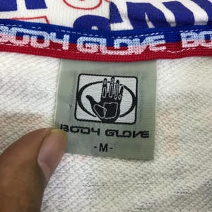 以下が含まれることがあります： 白いスウェットシャツのタグのクローズアップ。Body Gloveのロゴ、楕円の中の手。タグには「BODY GLOVE」とサイズ「-M-」と記載されています。スウェットシャツの上部には青と赤の文字があります。