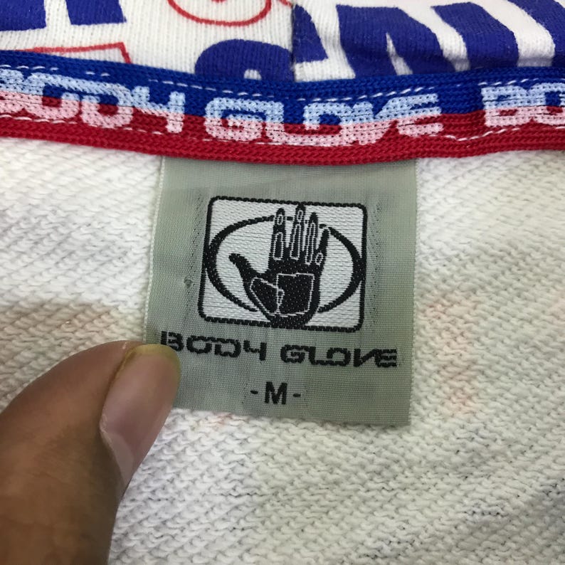 以下が含まれることがあります： 白いスウェットシャツのタグのクローズアップ。Body Gloveのロゴ、楕円の中の手。タグには「BODY GLOVE」とサイズ「-M-」と記載されています。スウェットシャツの上部には青と赤の文字があります。
