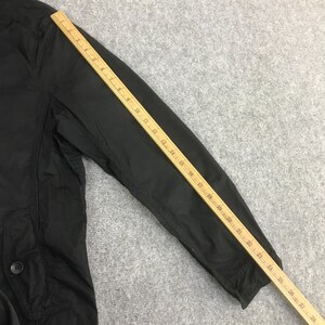 May include: A black jacket sleeve with a visible pocket featuring a single button closure. A wooden ruler is placed diagonally across the sleeve, showing a measurement length extending from approximately 1 inch to 28 inches along the arm.