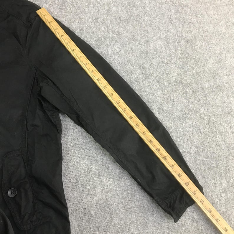 May include: A black jacket sleeve with a visible pocket featuring a single button closure. A wooden ruler is placed diagonally across the sleeve, showing a measurement length extending from approximately 1 inch to 28 inches along the arm.