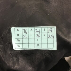 Może przedstawiać: Zbliżenie etykiety z ręcznie napisanymi cyframi i literami. Etykieta jest biała z siatką p&oacute;l. Numery obejmują 76, 21, 19, 26, 25 i 2. Tło to ciemna, teksturowana tkanina.