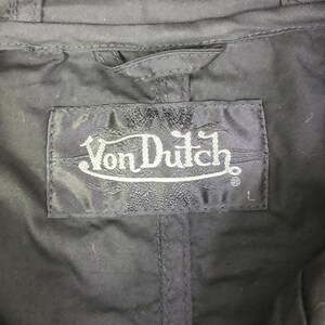 May include: A black garment featuring a rectangular fabric label stitched at the neckline. The label displays the white embroidered text Von Dutch in a stylized font, with the letter L positioned in the bottom right corner of the label.
