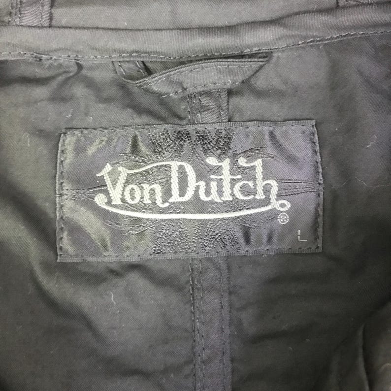 May include: A black garment featuring a rectangular fabric label stitched at the neckline. The label displays the white embroidered text Von Dutch in a stylized font, with the letter L positioned in the bottom right corner of the label.
