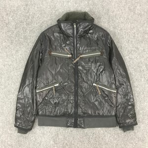 Może przedstawiać: Ciemnoszara pikowana kurtka z wysokim kołnierzem i wieloma kieszeniami zapinanymi na zamek. Kurtka posiada zamek błyskawiczny na całej długości oraz prążkowany d&oacute;ł i mankiety. Kurtka ma pikowany wz&oacute;r i miękki, teksturowany kołnierz. Kurtka ma napis na piersi.