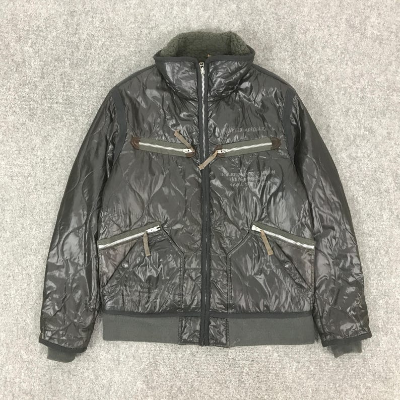 Może przedstawiać: Ciemnoszara pikowana kurtka z wysokim kołnierzem i wieloma kieszeniami zapinanymi na zamek. Kurtka posiada zamek błyskawiczny na całej długości oraz prążkowany d&oacute;ł i mankiety. Kurtka ma pikowany wz&oacute;r i miękki, teksturowany kołnierz. Kurtka ma napis na piersi.