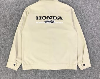 May include: A beige jacket with the Honda logo and "Mugen 無限 power" printed in black on the back. The jacket has a button-down front, two front pockets, and buttoned cuffs. The jacket is made of a durable material.