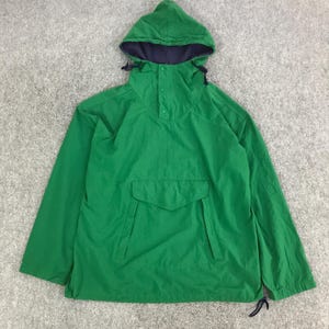 May include: A green anorak jacket with a hood and a front pocket. The hood has a navy blue lining and drawstrings. The jacket has a button-up closure and long sleeves. The jacket is made of a lightweight material.
