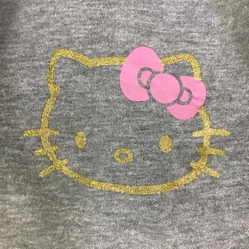 May include: Close-up of a gray fabric with a gold glitter outline of a cartoon cat face. The cat has a pink bow and gold glitter eyes and nose. The design is centered on the fabric.