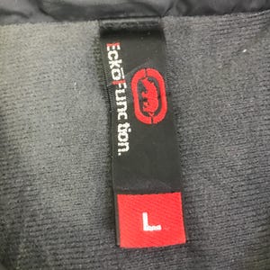 May include: A black fabric clothing tag featuring the white text Ecko Unltd. Function and a red rhinoceros logo. Below the logo, a separate red rectangular section displays the white letter L, indicating a size large, attached to a dark gray fabric.