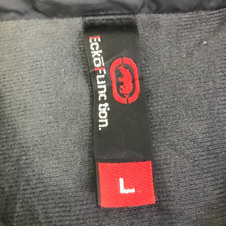 May include: A black fabric clothing tag featuring the white text Ecko Unltd. Function and a red rhinoceros logo. Below the logo, a separate red rectangular section displays the white letter L, indicating a size large, attached to a dark gray fabric.