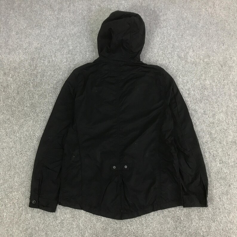 May include: A solid black hooded jacket featuring long sleeves with buttoned cuffs, a full-length front zipper, and a small horizontal tab detail with two buttons positioned at the lower center back of the garment, providing a structured appearance.
