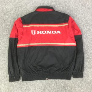May include: A black and red zip-up jacket featuring the Honda logo and name in white text across the back. The garment includes horizontal beige stripes across the chest and back, a fold-down collar, and elasticized cuffs and hem for a secure fit.