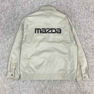 Op de afbeelding: Een lichtgrijze jas met het zwarte "MAZDA" logo op de rug. De jas heeft een knoopsluiting aan de voorkant, twee borstzakken en manchetten met knopen. De jas is gemaakt van een lichtgewicht materiaal.