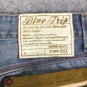 K&ouml;nnte beinhalten: Nahaufnahme einer hellblauen Jeans mit einem Lederaufn&auml;her im Used-Look. Der Aufn&auml;her tr&auml;gt die Aufschrift "Blue Trip" und "Edwin 503" mit zus&auml;tzlichem Text. Die Jeans hat eine braune Wildleder-Tasche.