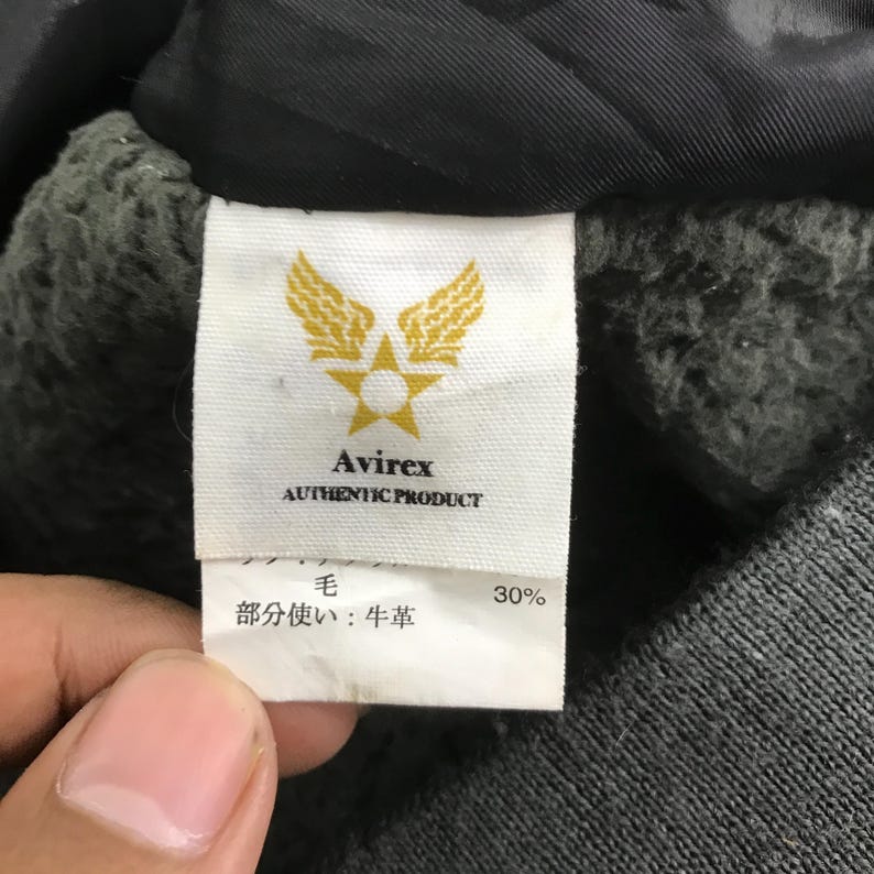 Może przedstawiać: Zbliżenie na metkę produktu Avirex. Etykieta jest biała ze złotymi literami i złotym emblematem skrzydeł i gwiazdy. Tekst na etykiecie brzmi "Avirex AUTHENTIC PRODUCT". Na etykiecie znajduje się r&oacute;wnież japoński tekst i liczba 30%.