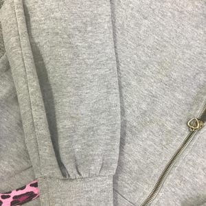 May include: Close-up of a gray sweatshirt with a zipper and a heart-shaped zipper pull. The sleeve cuff has a pink and black patterned design. The fabric appears to be a soft, textured material.