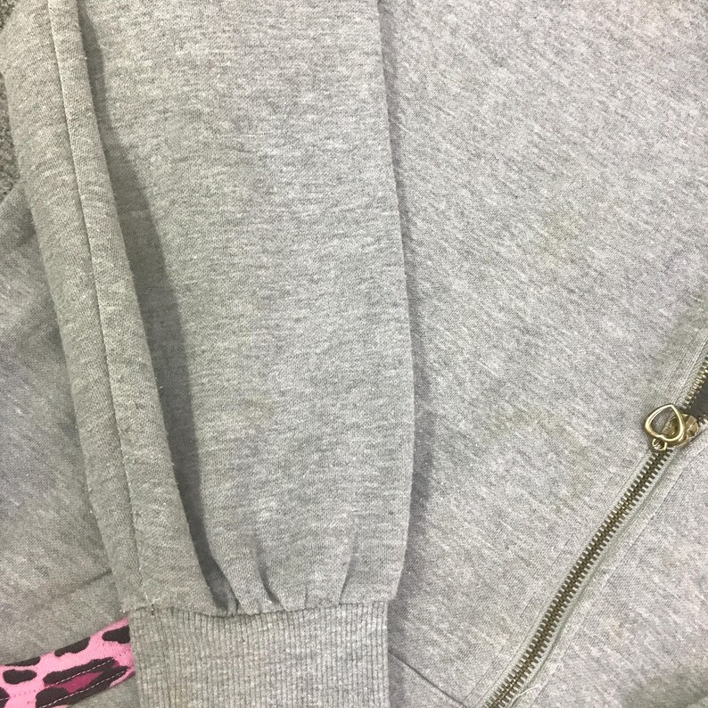 May include: Close-up of a gray sweatshirt with a zipper and a heart-shaped zipper pull. The sleeve cuff has a pink and black patterned design. The fabric appears to be a soft, textured material.