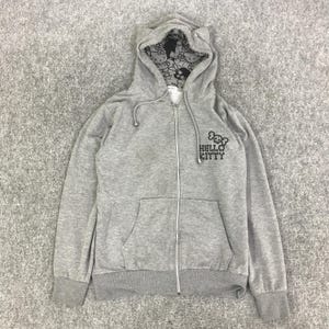 Może przedstawiać: Szara bluza z kapturem zapinana na zamek z logo Hello Kitty i wzorem czaszki i skrzyżowanych piszczeli. Wnętrze kaptura ma wz&oacute;r Hello Kitty. Bluza ma przednią kieszeń i sznurek. Zamek błyskawiczny jest srebrny.