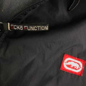 May include: A black jacket featuring a zipper pull with the text ECKO FUNCTION in white and red lettering. A small rectangular red patch is sewn onto the fabric, displaying a white graphic of a rhinoceros inside an oval outline logo.
