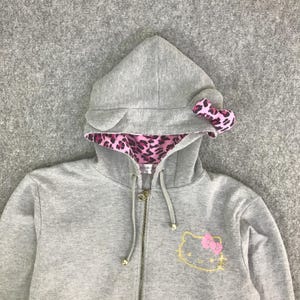 May include: A gray zip-up hoodie featuring a hood with attached ears and a pink leopard print lining. The hood has a pink bow. The front of the hoodie has a gold embroidered Hello Kitty design with a pink bow.