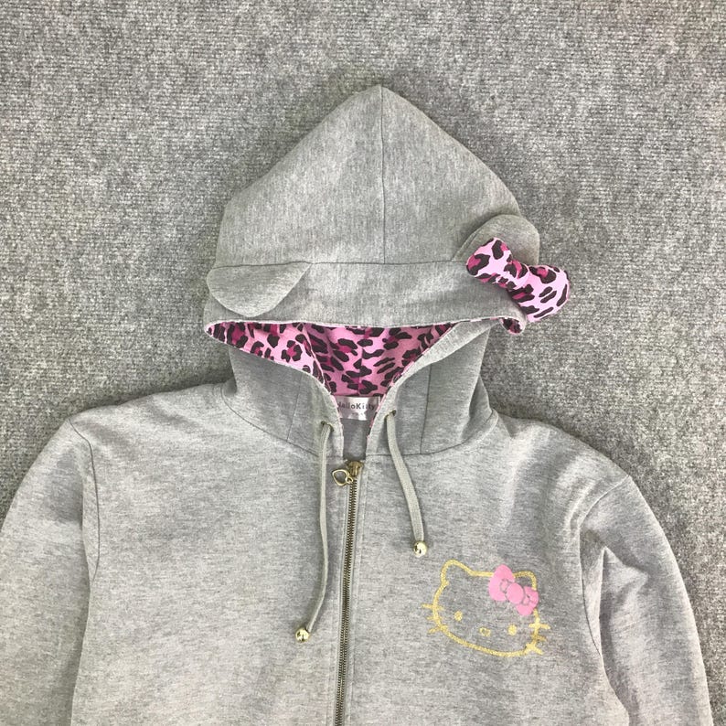 May include: A gray zip-up hoodie featuring a hood with attached ears and a pink leopard print lining. The hood has a pink bow. The front of the hoodie has a gold embroidered Hello Kitty design with a pink bow.