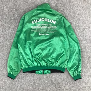 Puede incluir: Una chaqueta de satén verde con cremallera negra y cintura negra. La parte posterior de la chaqueta tiene un logotipo blanco que dice "FUJICOLOR Película altamente avanzada para impresiones en color absolutamente brillantes. Por fin, una película que realmente te convierte en un mejor fotógrafo. FUJI FILM".