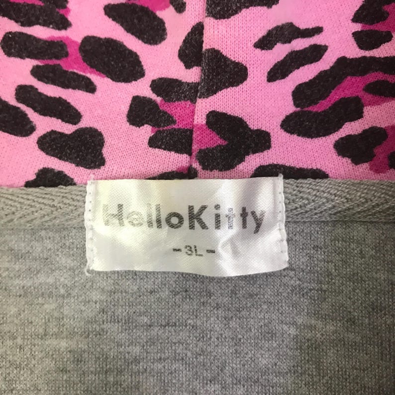 May include: Close-up of a gray hoodie with a pink leopard print hood. The label reads "Hello Kitty" in white text. The leopard print features black spots on a pink background.