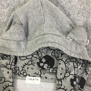Może przedstawiać: Szara bluza z kapturem z podszewką we wz&oacute;r Hello Kitty i czaszki w kolorze czarno-białym. Kaptur ma małe detale w kształcie uszu i metalowy suwak. Etykieta wewnątrz bluzy głosi "Hello Kitty".