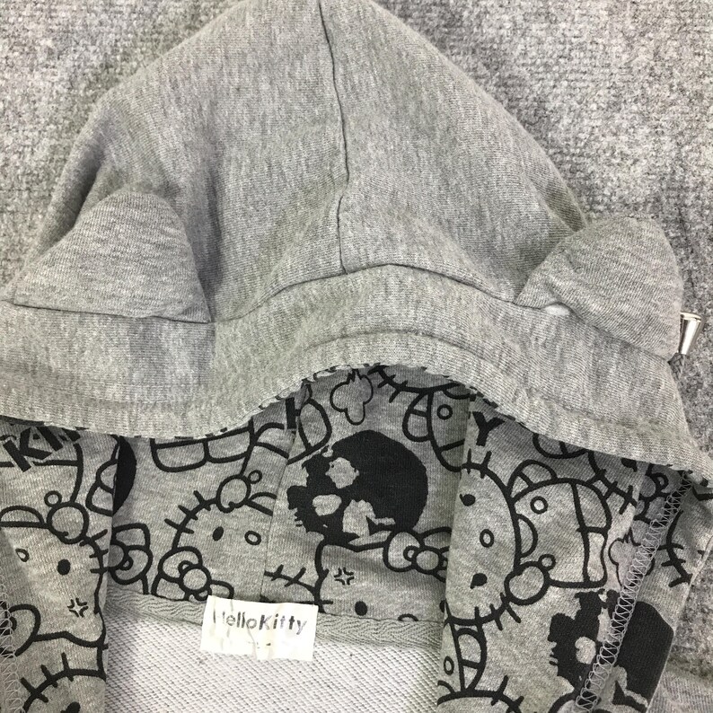 Może przedstawiać: Szara bluza z kapturem z podszewką we wz&oacute;r Hello Kitty i czaszki w kolorze czarno-białym. Kaptur ma małe detale w kształcie uszu i metalowy suwak. Etykieta wewnątrz bluzy głosi "Hello Kitty".