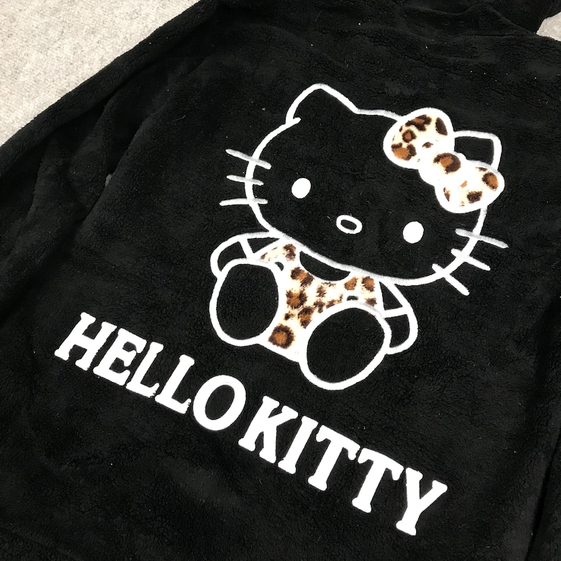 Pu&ograve; includere: Felpa nera in pile con un grafico bianco di Hello Kitty. Il grafico presenta Hello Kitty con un outfit a stampa leopardata e un fiocco.