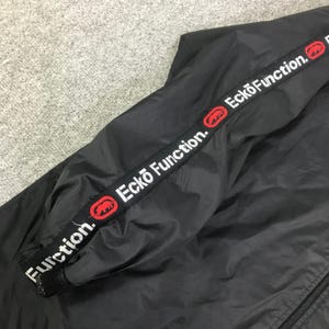 May include: A black windbreaker jacket featuring a prominent black fabric strip running along the sleeve. The strip displays the white text Ecko Function and a red rhinoceros logo repeated multiple times throughout the length of the band.