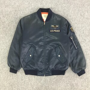 Op de afbeelding: Marineblauwe bomberjack met ritssluiting, geribde manchetten en kraag. Voorzien van een "POLICE U.S. POLICE" patch op de borst en een patch op de mouw. De jas heeft twee voorzakken en een zak met rits op de linkermouw.