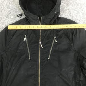 May include: A black hooded jacket with a central zipper and two diagonal zippered chest pockets. A yellow ruler is placed horizontally across the chest, showing a width measurement of approximately 18 inches from the left to the right side seam.