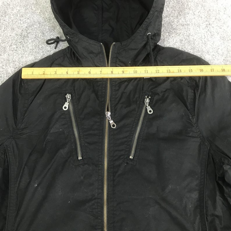 May include: A black hooded jacket with a central zipper and two diagonal zippered chest pockets. A yellow ruler is placed horizontally across the chest, showing a width measurement of approximately 18 inches from the left to the right side seam.
