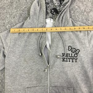 Może przedstawiać: Szara bluza z kapturem zapinana na zamek z motywem czaszki Hello Kitty. Bluza posiada gwiazdkowy suwak i wzorzystą podszewkę kaptura. Linijka pokazuje wymiar około 43 cm.