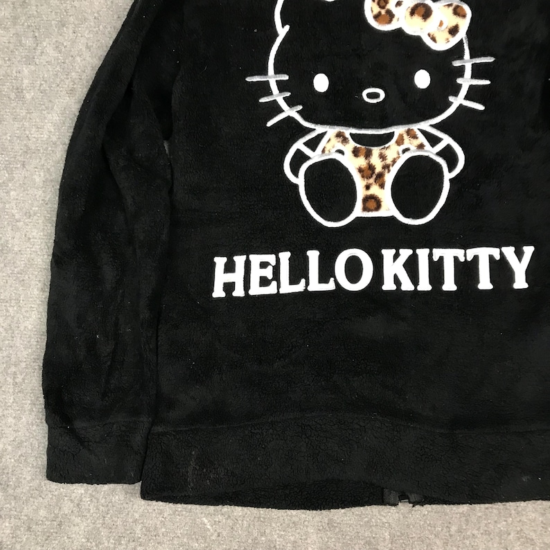 Pu&ograve; includere: Giacca in pile nera con una grafica bianca di Hello Kitty e un design a stampa leopardata. La giacca ha una chiusura a zip e maniche lunghe.