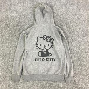 Może przedstawiać: Szara bluza z kapturem z czarną grafiką Hello Kitty, w tym czaszką i skrzyżowanymi piszczelami. Poniżej nadrukowano napis "HELLO KITTY". Bluza posiada kaptur ze sznurkiem oraz prążkowane mankiety i d&oacute;ł. Wygodny, codzienny element garderoby.