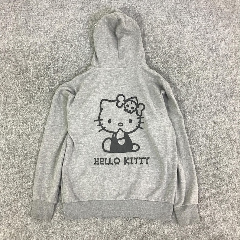 Może przedstawiać: Szara bluza z kapturem z czarną grafiką Hello Kitty, w tym czaszką i skrzyżowanymi piszczelami. Poniżej nadrukowano napis "HELLO KITTY". Bluza posiada kaptur ze sznurkiem oraz prążkowane mankiety i d&oacute;ł. Wygodny, codzienny element garderoby.