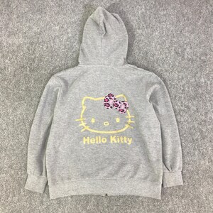 May include: A gray zip-up hoodie featuring a Hello Kitty graphic on the back. The graphic includes a yellow outline of Hello Kitty's face, a pink and purple bow, and the words "Hello Kitty" in yellow.