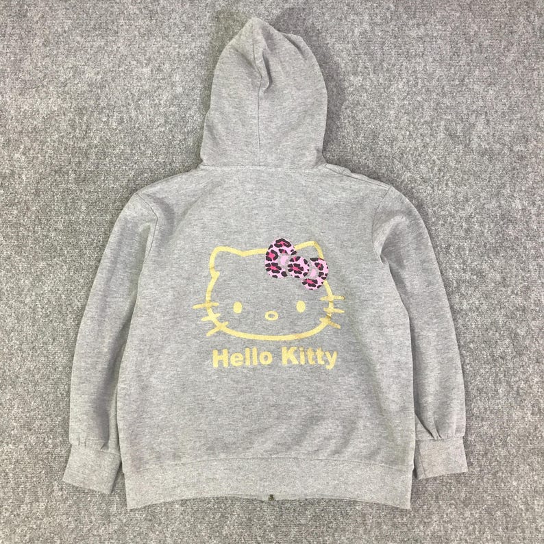 May include: A gray zip-up hoodie featuring a Hello Kitty graphic on the back. The graphic includes a yellow outline of Hello Kitty's face, a pink and purple bow, and the words "Hello Kitty" in yellow.