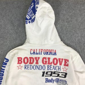 以下が含まれることがあります： 「CALIFORNIA BODY GLOVE REDONDO BEACH 1953」と書かれた青と赤の文字が入った白いパーカー。フードには「CALIFORNIA BODY GLOVE」と書かれた青い文字が入っています。「CALIFORNIA」の文字も左側に縦にプリントされています。